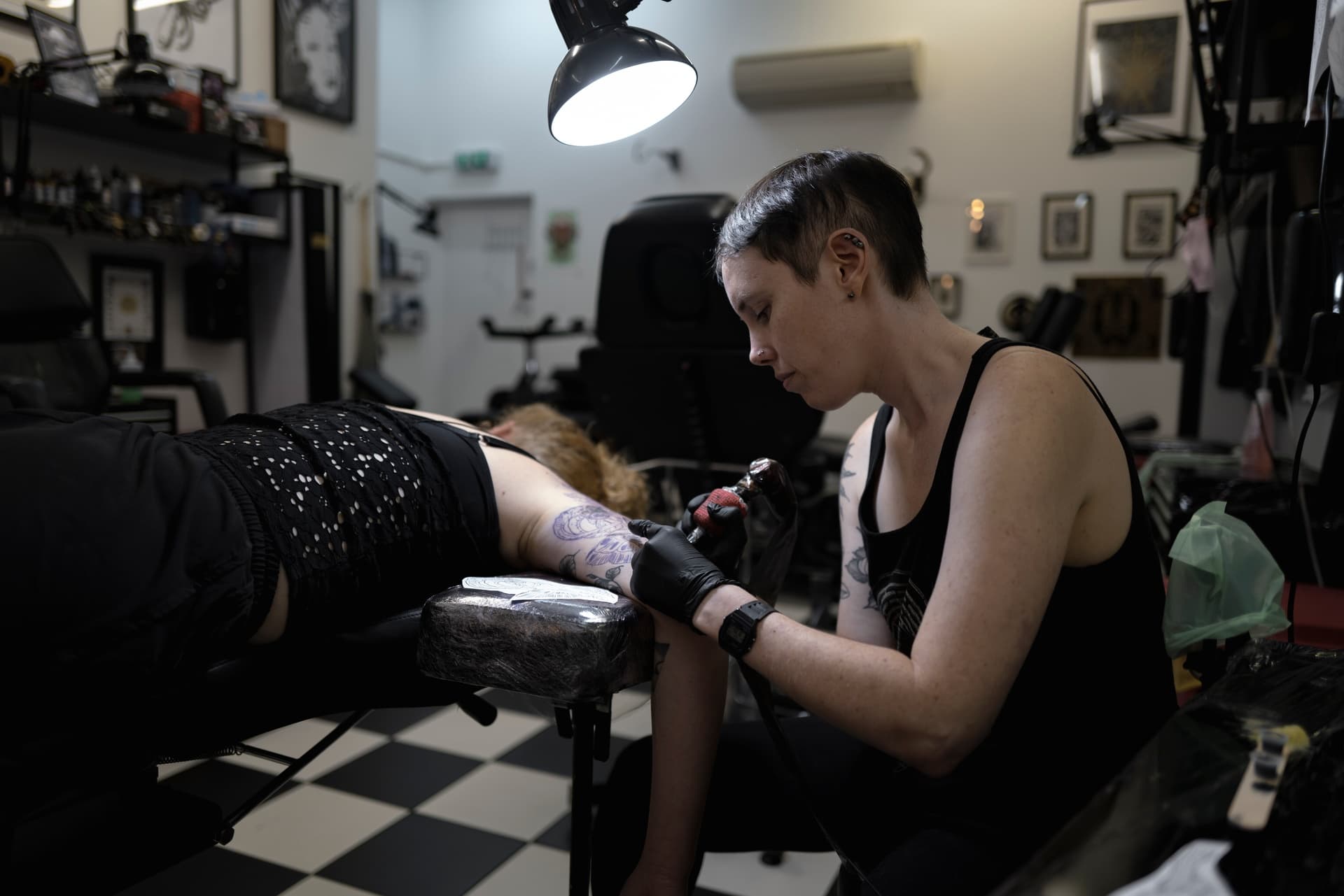 A tattoo artist with short dark hair and tattoos on their arms tattooing a person's arm in a tattoo studio with black and white checkered floors, framed pictures on the wall, and various tattoo equipment around.