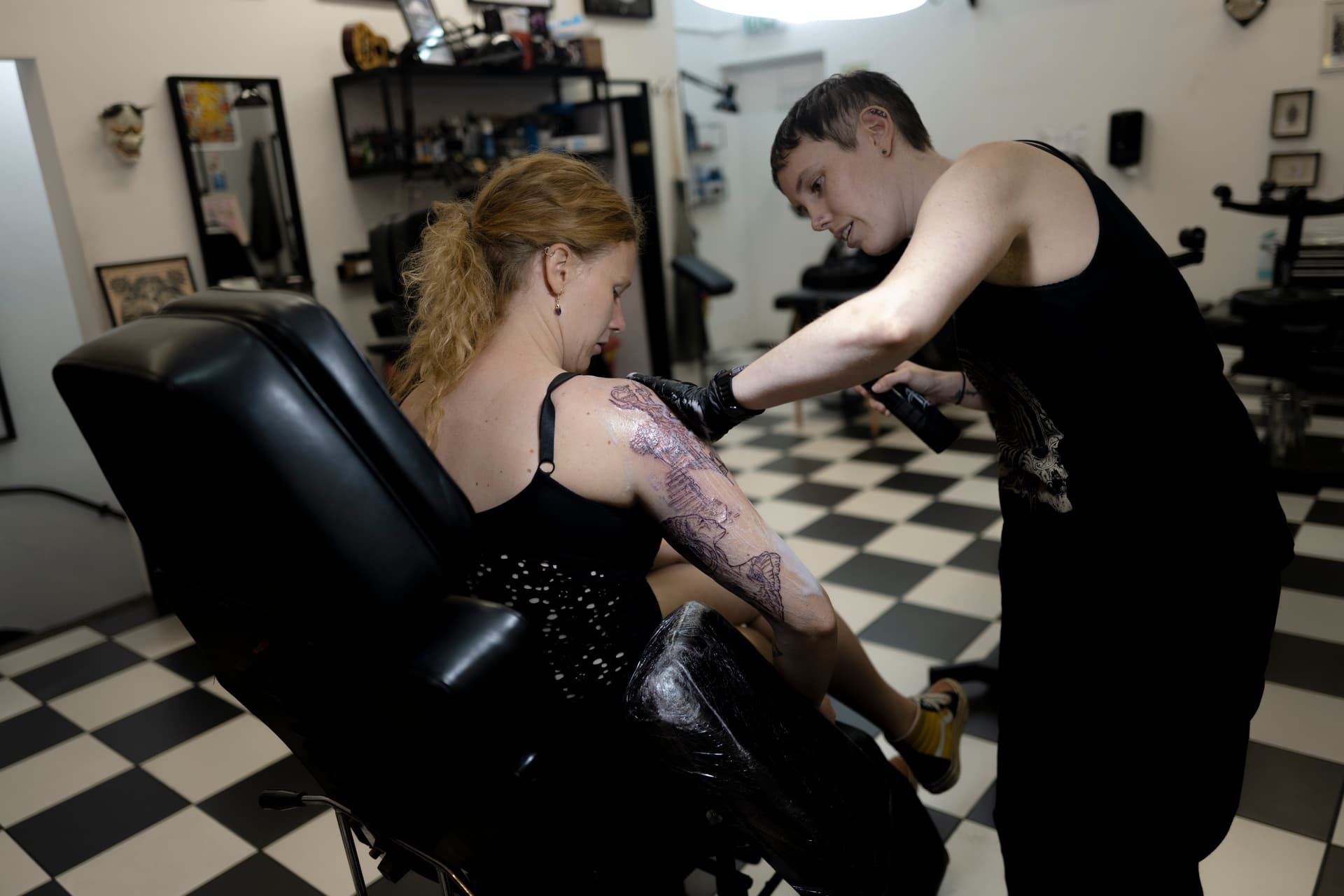 A woman with curly hair gets a tattoo on her arm while seated in a tattoo parlor. She is being assisted by a tattoo artist who is wearing black gloves.