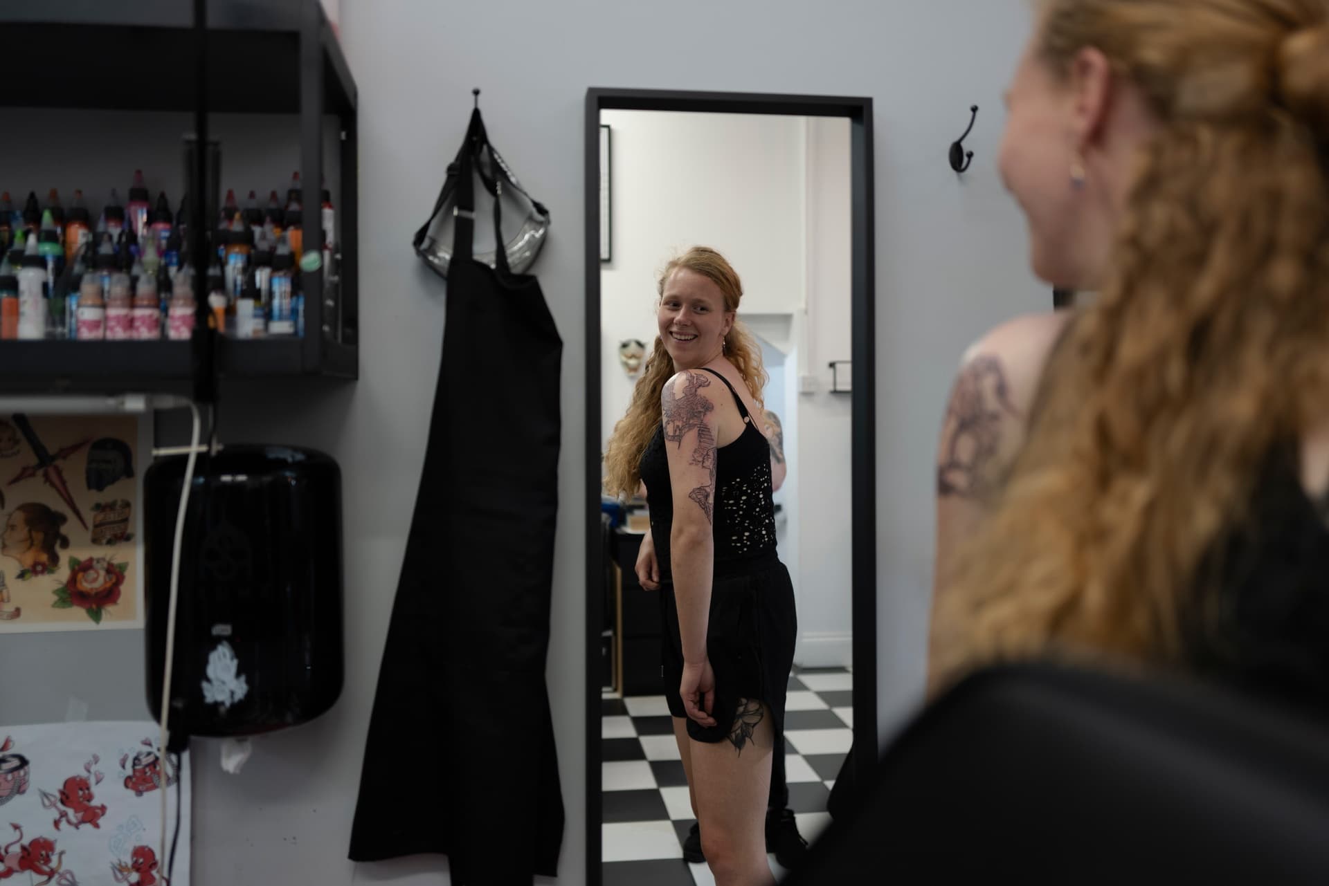 A woman with long curly red hair looking at herself in a full-length mirror, smiling, with tattoos on her arm and leg, in a tattoo or beauty salon.