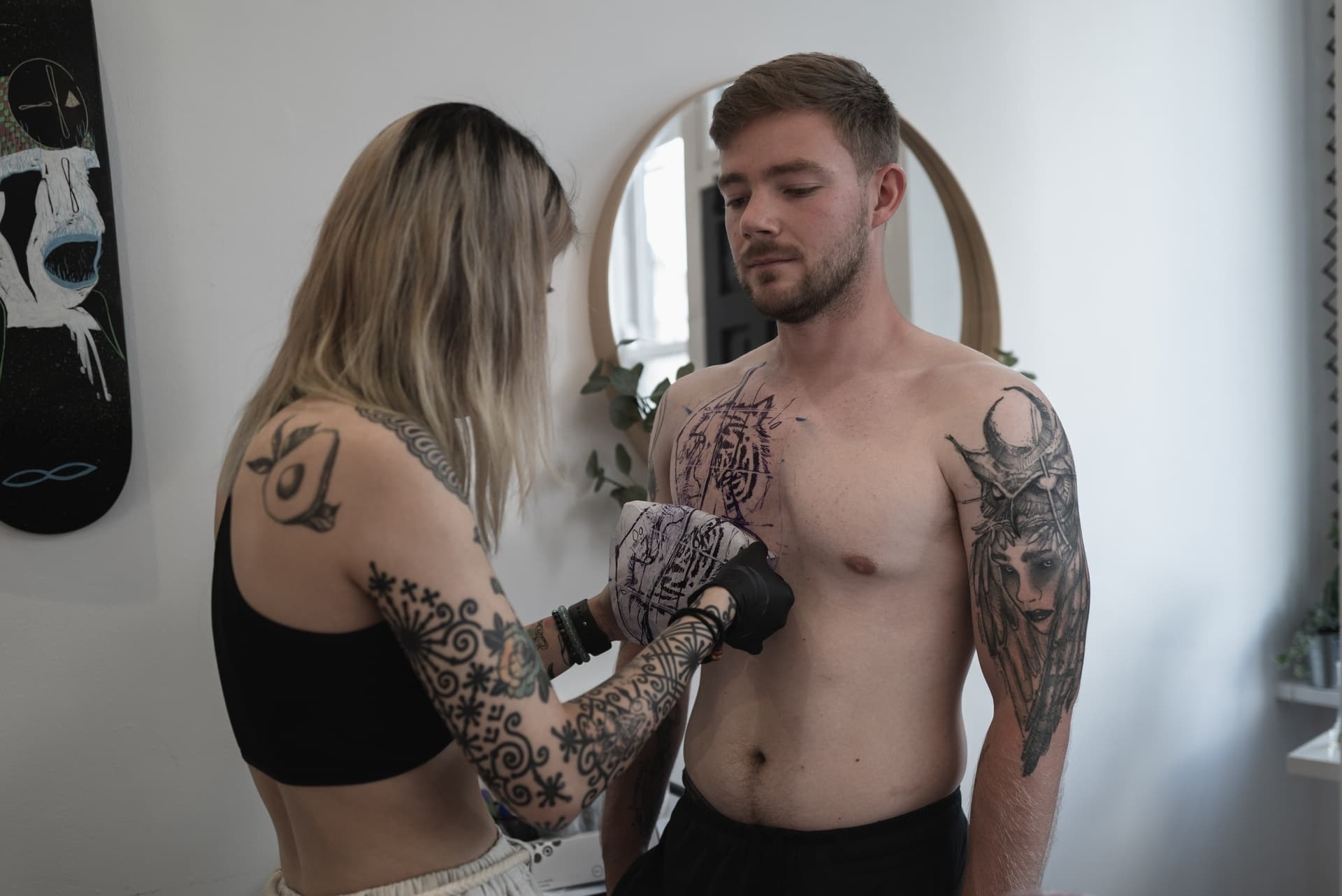 Tattoo artist inks a tattoo on a shirtless man with tattooed arms in a studio with a mirror and artwork on the wall.