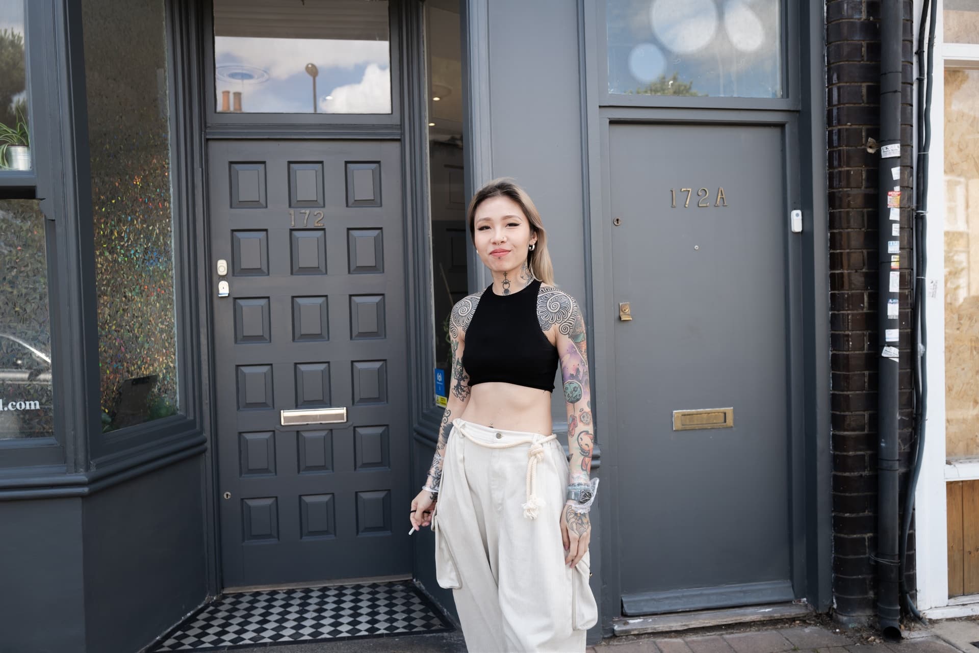 A person with tattoos, wearing a black crop top and white pants, stands outside a gray building with numbers 172 and 172A on the doors.