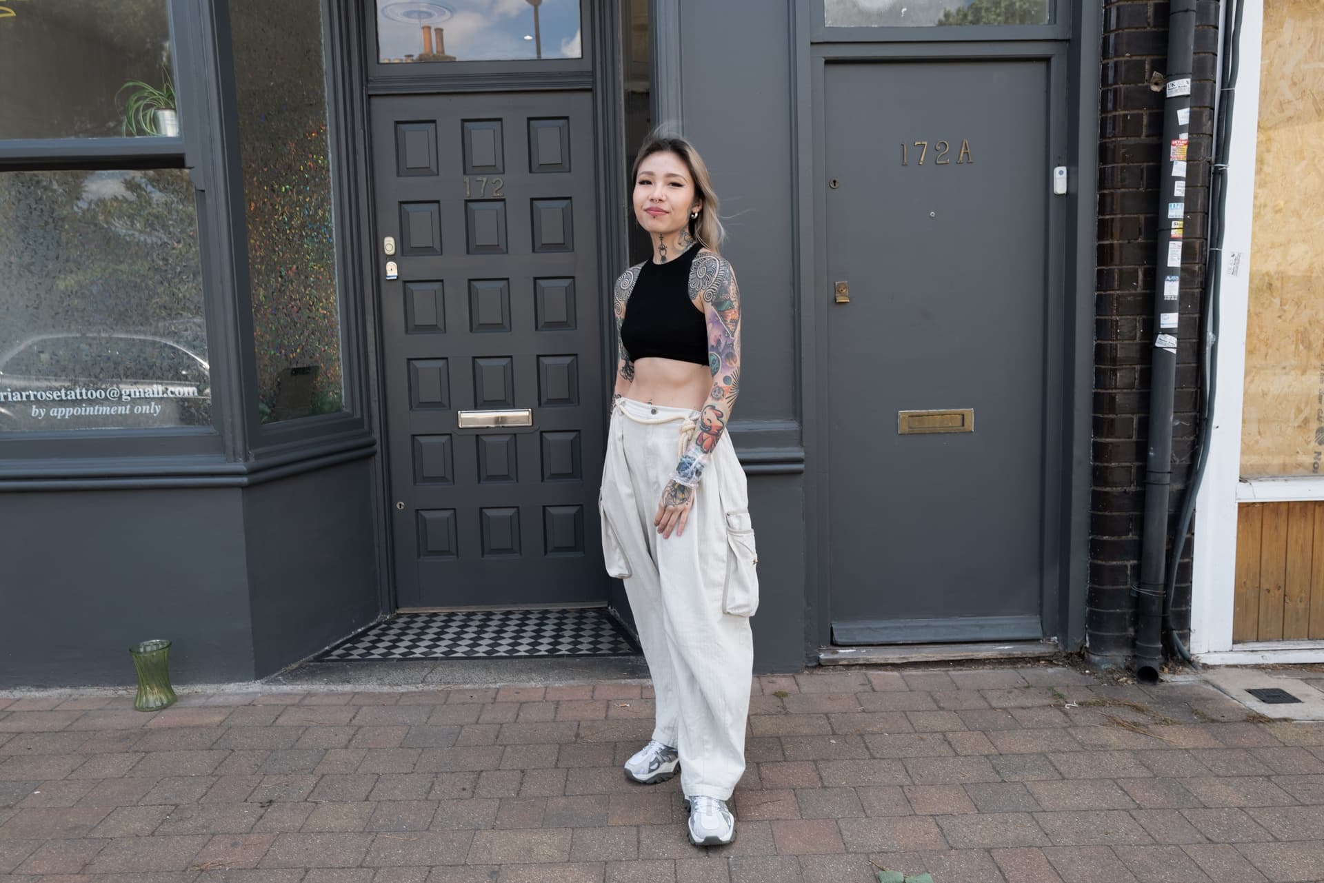 Young woman with tattoos in a black crop top and white cargo pants standing outside a building with a dark gray door and window, on a brick sidewalk.