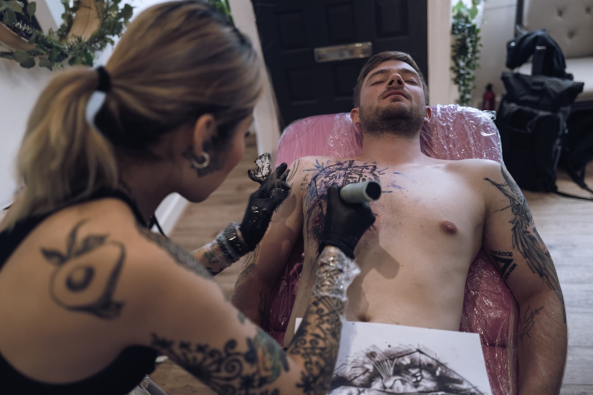 Tattoo artist creating a tattoo on a shirtless man lying on a chair, with the artist holding tattoo machine, on a wooden floor in a studio.