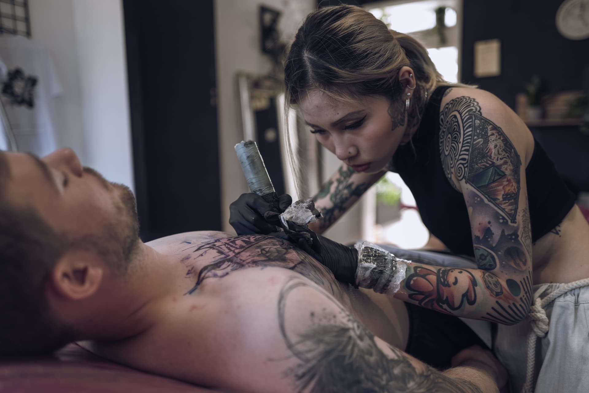 A woman with tattoos is tattooing a man's chest in a tattoo studio.
