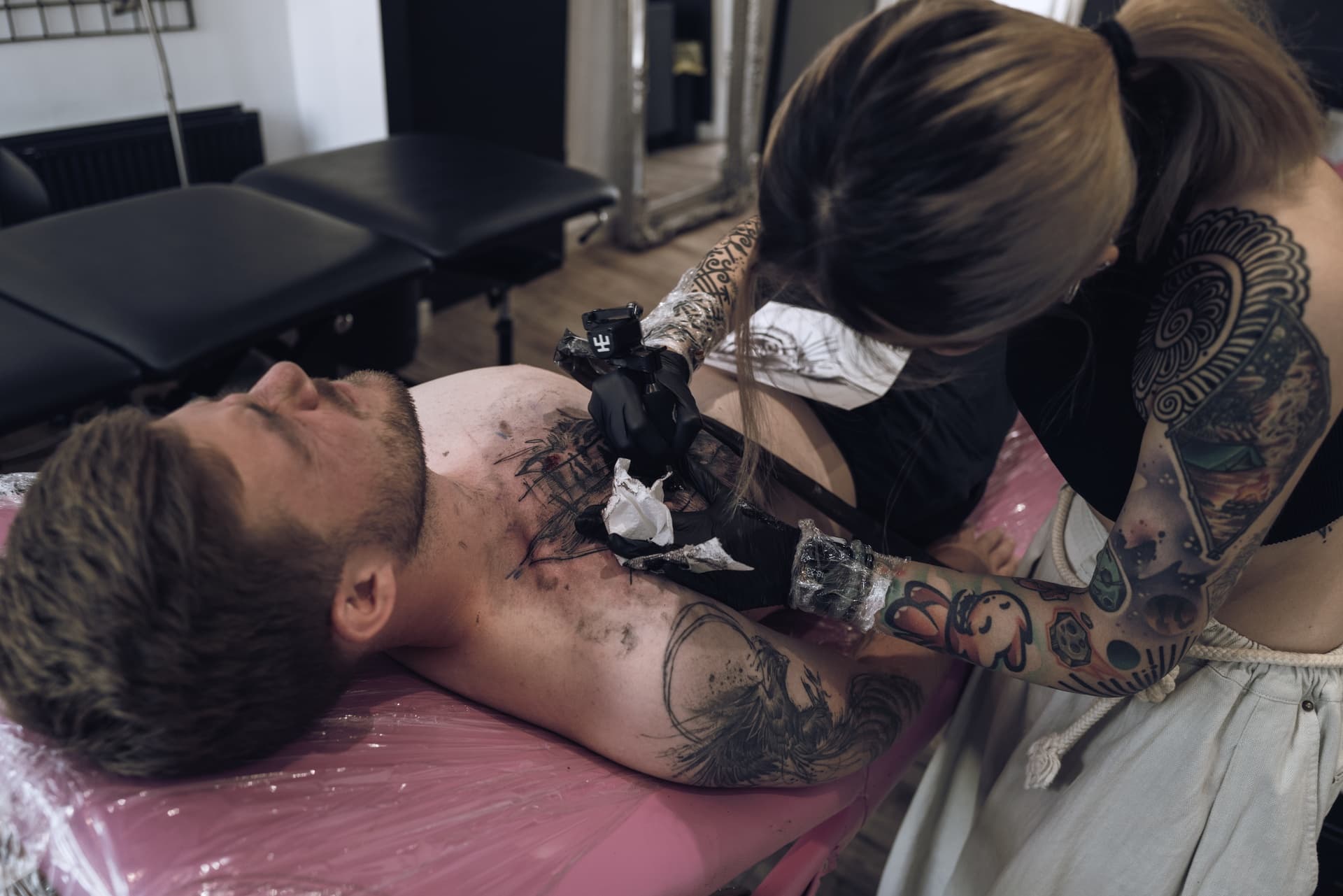 A woman with tattoos on her arms is tattooing a man's chest, who is lying on a tattooing bed with his eyes closed. The woman is wearing gloves and holding a tattoo machine, working on the man's chest, which already has some tattoo outlines.