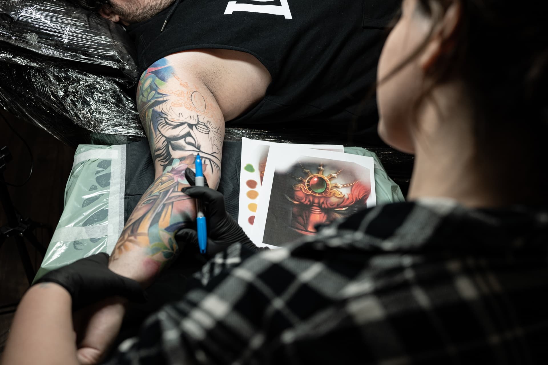 A tattoo artist working on a colorful tattoo on a client's forearm. The artist is wearing black gloves and using a tattoo machine. There are reference sheets with fantasy-themed artwork on the table.