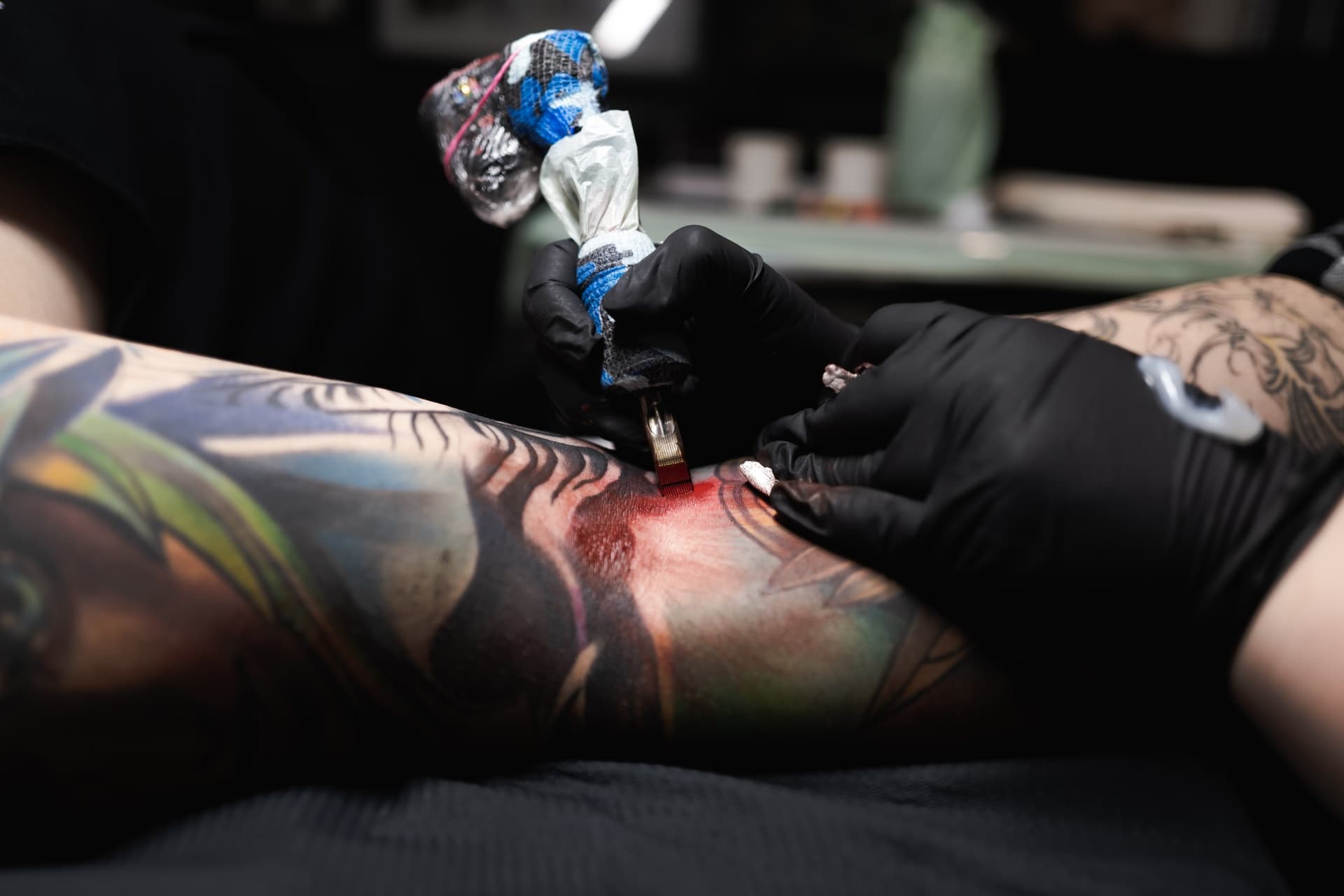 A tattoo artist wearing black gloves is tattooing a colorful tattoo on a person's arm with tattoos. The artist is using a tattoo machine, and the tattoo is in progress.