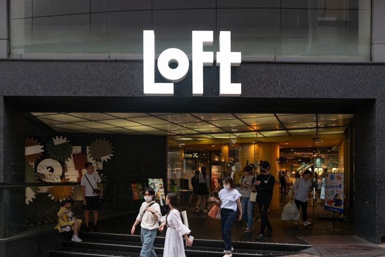 People exiting the LOFT store with large white sign above entrance, glass doors, and interior visible, in an urban setting.