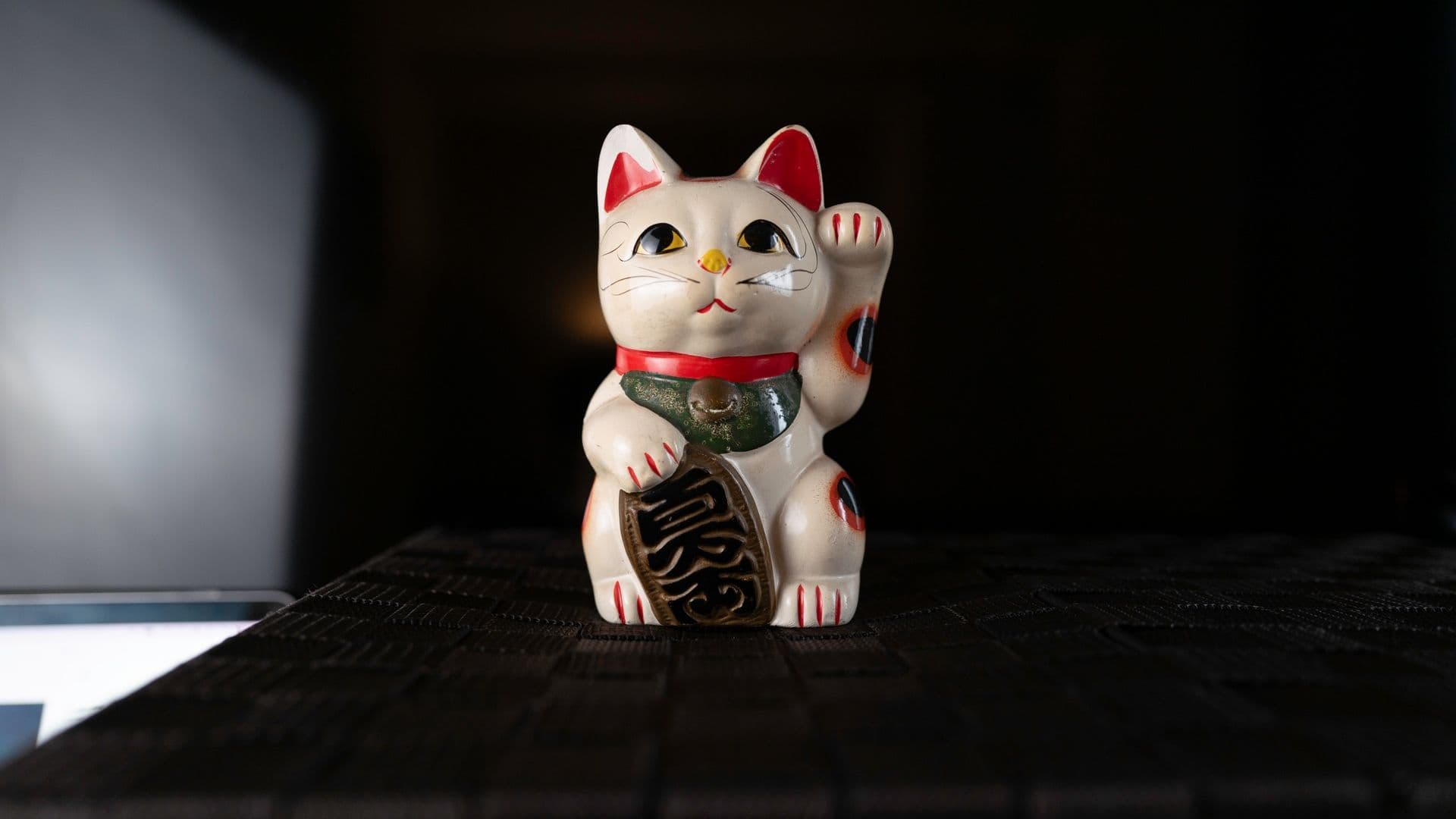 Maneki-neko, a Japanese ceramic cat figurine with a raised paw, sitting on a textured black surface, against a dark background.