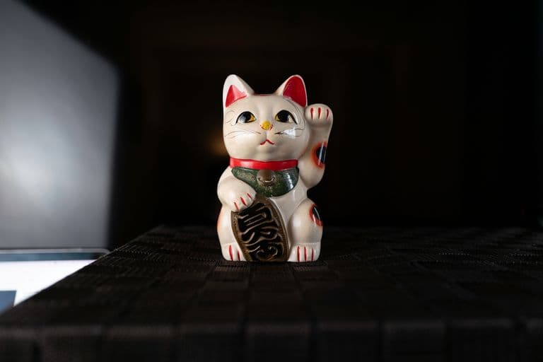 A ceramic Maneki-neko, or Japanese lucky cat, with its right paw raised, sitting on a black textured surface, with a dark background and a partial view of a silver laptop on the left