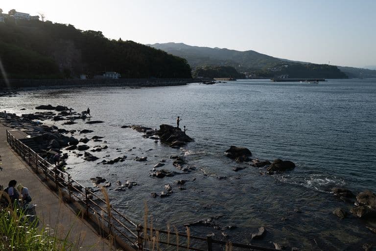 People fishing on rocks along a waterfront with a pathway and railing in the foreground, mountains and a bay in the background.