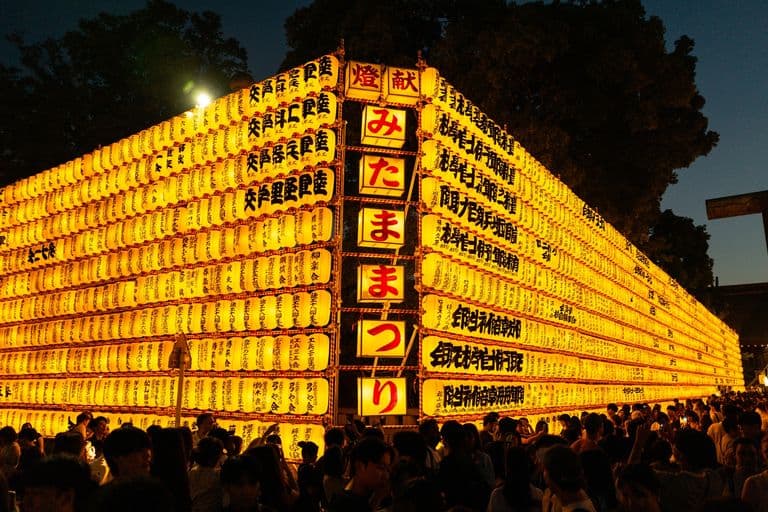 A large display of hanging illuminated yellow paper lanterns with Japanese characters written on them, with a crowd of people gathered underneath at night.