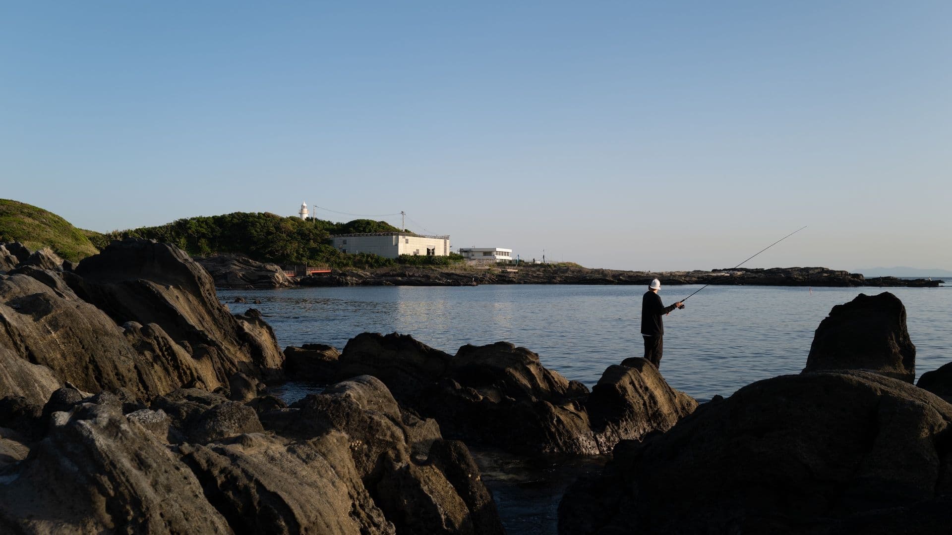 A person fishing on rocks by the seaside during sunset, with calm water and a distant white building with a lighthouse in the background.