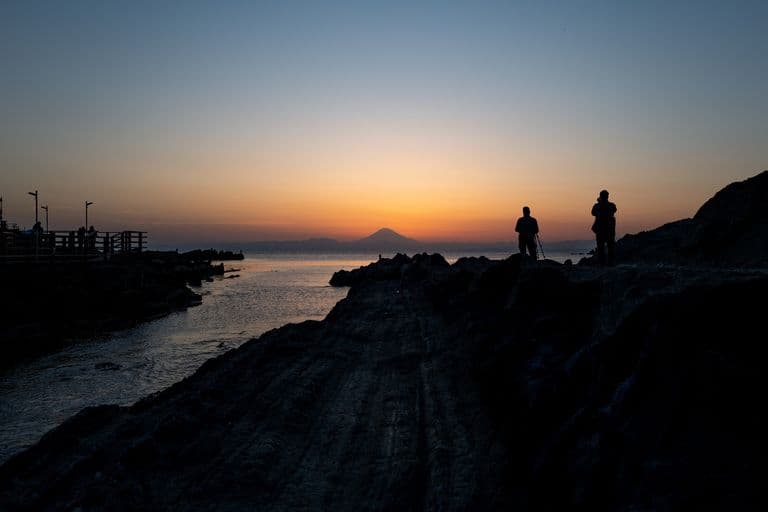 Silhouettes of two people standing on a rocky shoreline at sunset, with one person taking a photo and the other holding a camera, overlooking a body of water with distant mountains, including Mount Fuji.