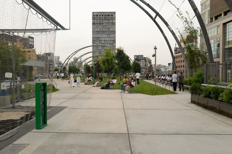People sitting on benches and walking in a city park with modern buildings and trees, under a cloudy sky.