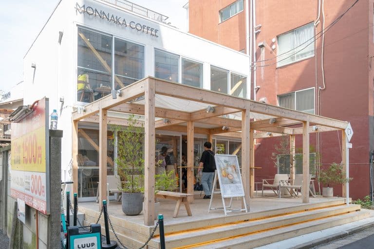Exterior of Monnaka Coffee shop with wooden deck and seating area, people entering, and building with large windows behind.
