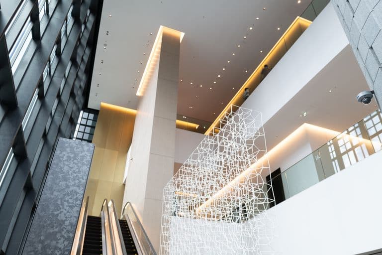 Interior of a modern building with escalator, hanging geometric art installation, and multiple levels with people walking.