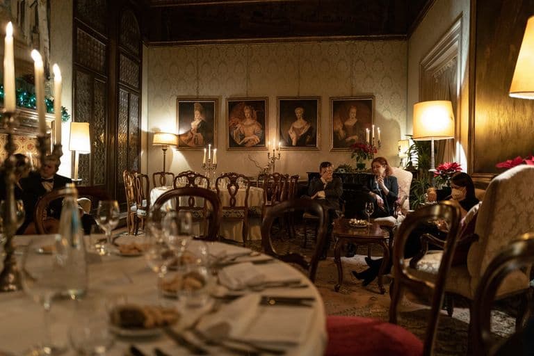 Elegant dining room with dim lighting, decorated for a festive occasion, with empty tables set with glassware and plates, and people sitting and talking in armchairs, wearing face masks.