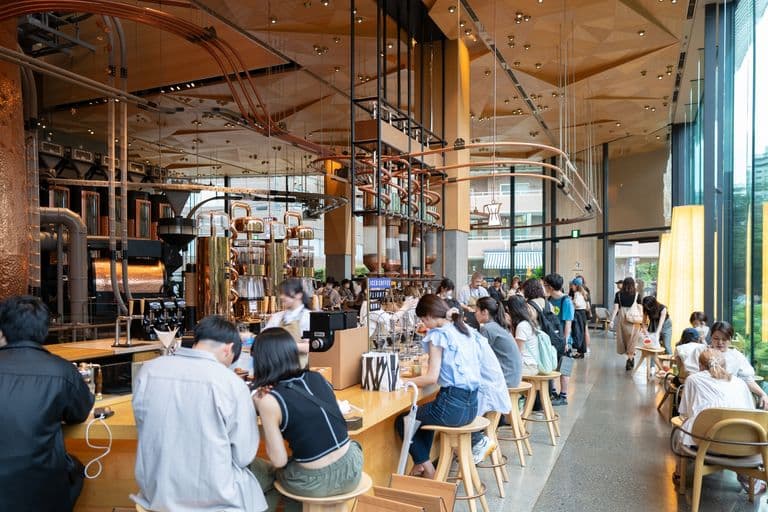 Inside a busy modern coffee shop with a long counter, baristas preparing drinks, and customers sitting on stools and chairs enjoying their beverages. Large windows let in natural light, and intricate industrial-style piping decor is visible on the ceiling.
