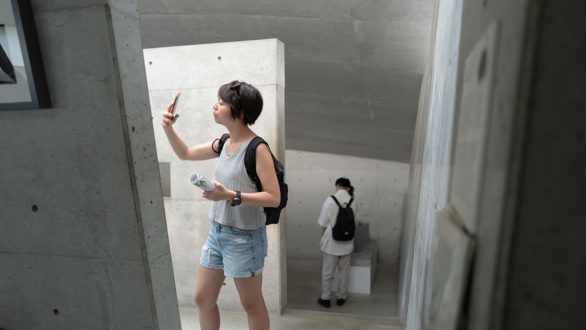 A young woman taking a selfie with her smartphone inside a modern concrete building, with another person standing in the background on the stairs.