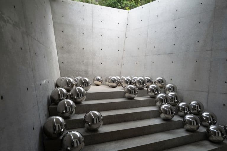 Concrete staircase surrounded by bare concrete walls with numerous shiny metal spheres scattered across the steps and floor, reflecting the surrounding space.