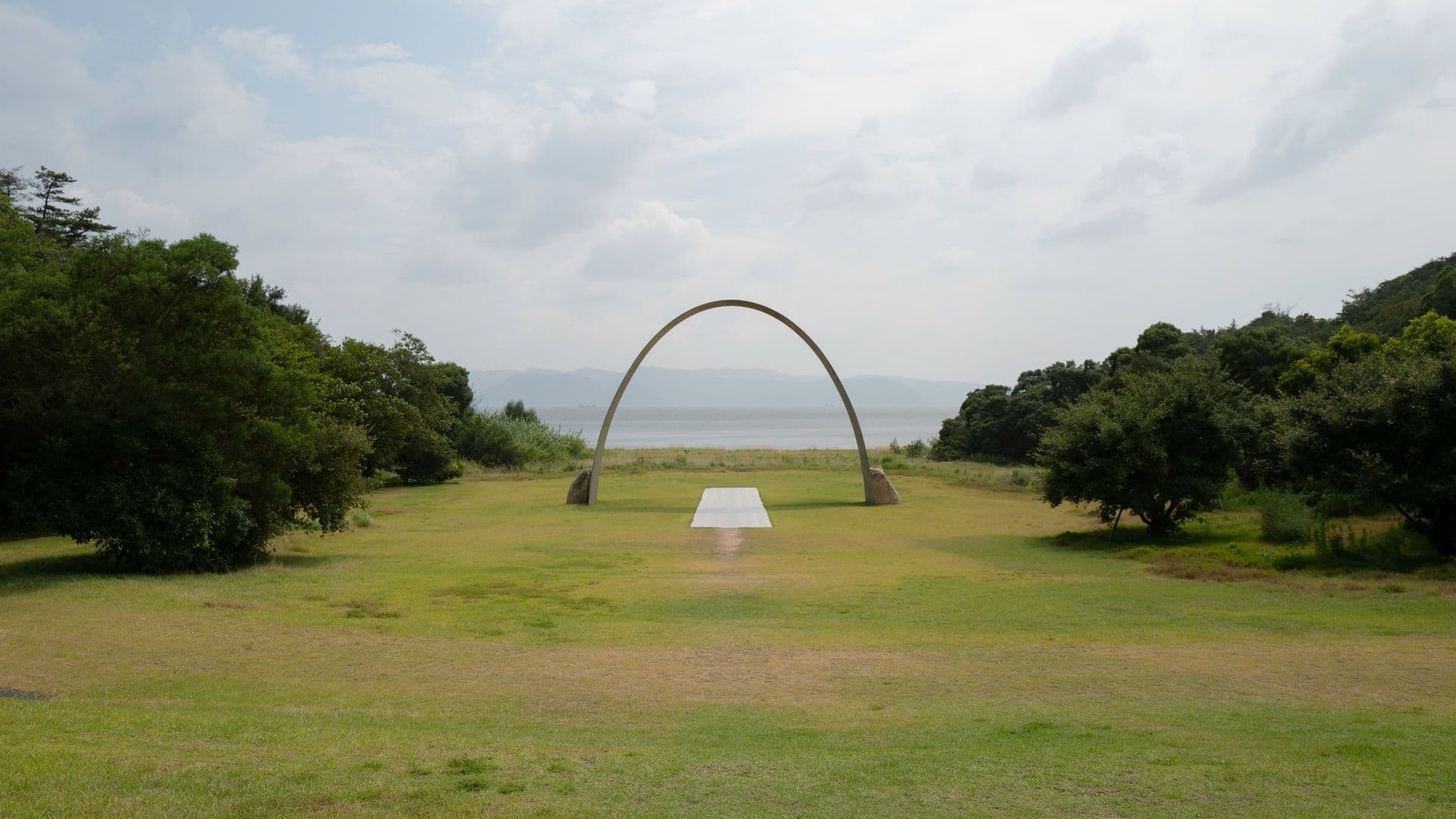 A grassy field with a large concrete arch and a white platform leading towards a body of water in the distance, surrounded by trees with mountains and cloudy sky in the background.