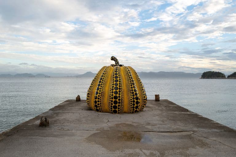A large pumpkin-shaped sculpture painted with yellow and black polka dots, situated on a concrete pier by the water, with islands and mountains in the distance under a cloudy sky.