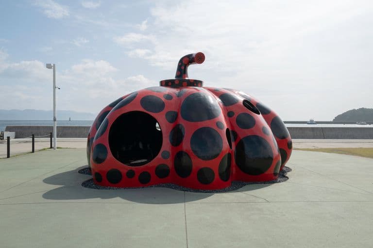 A playful, pumpkin-shaped sculpture painted with red and black spots, situated outdoors on a paved area near a body of water with landmasses in the background.