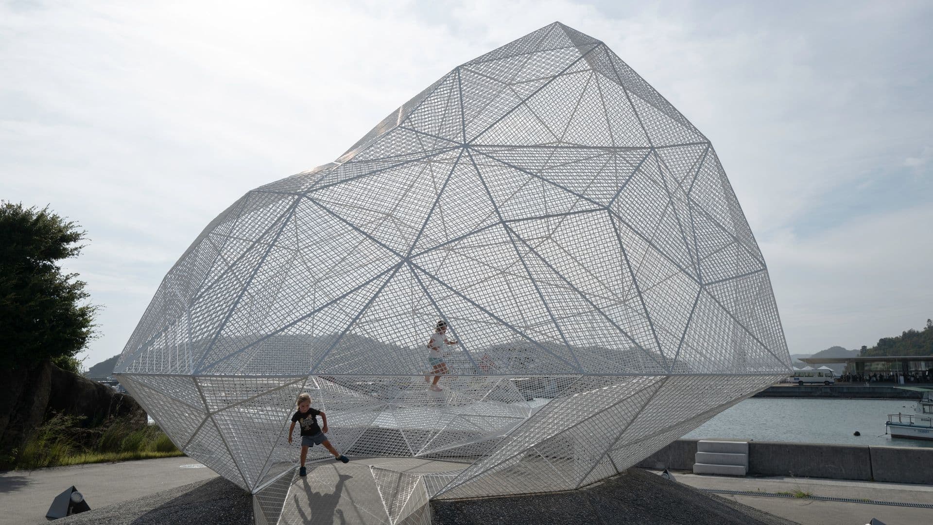 Large geometric metal sculpture resembling a crystalline structure with children playing inside, located outdoors near a body of water.