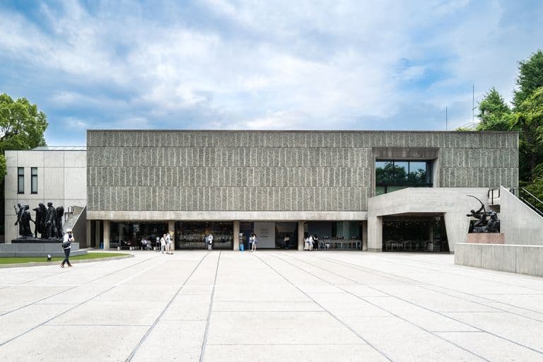 Modern museum with large entrance, sculptures on steps, and people entering and exiting in front, with trees and a partly cloudy sky in the background.