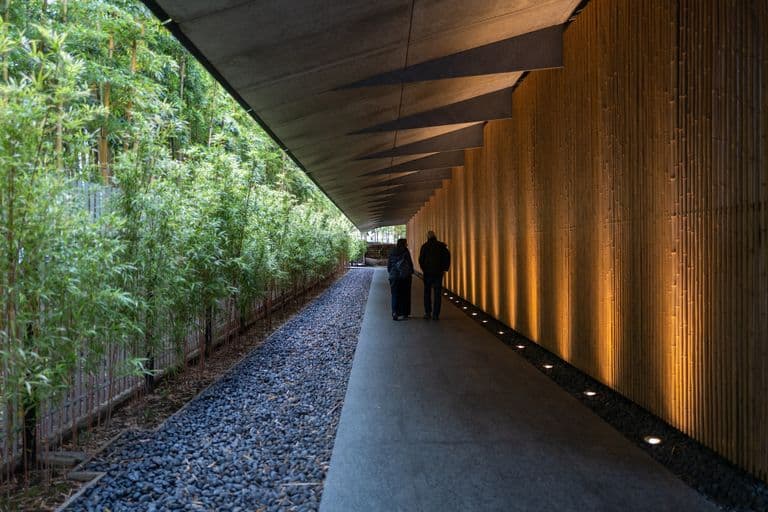 Three people walking in a modern, covered outdoor corridor with bamboo plants on one side and a wooden wall with warm lighting on the other.