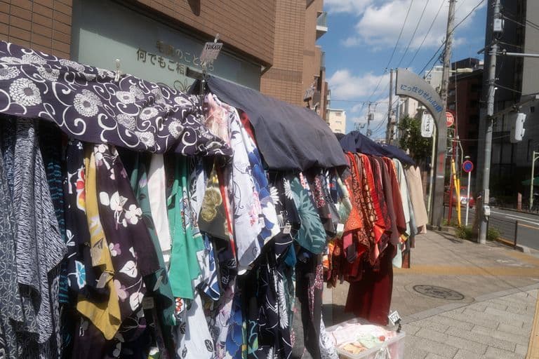 Outdoor clothes market with colorful Japanese yukata and summer garments hanging on racks under a sunny sky.