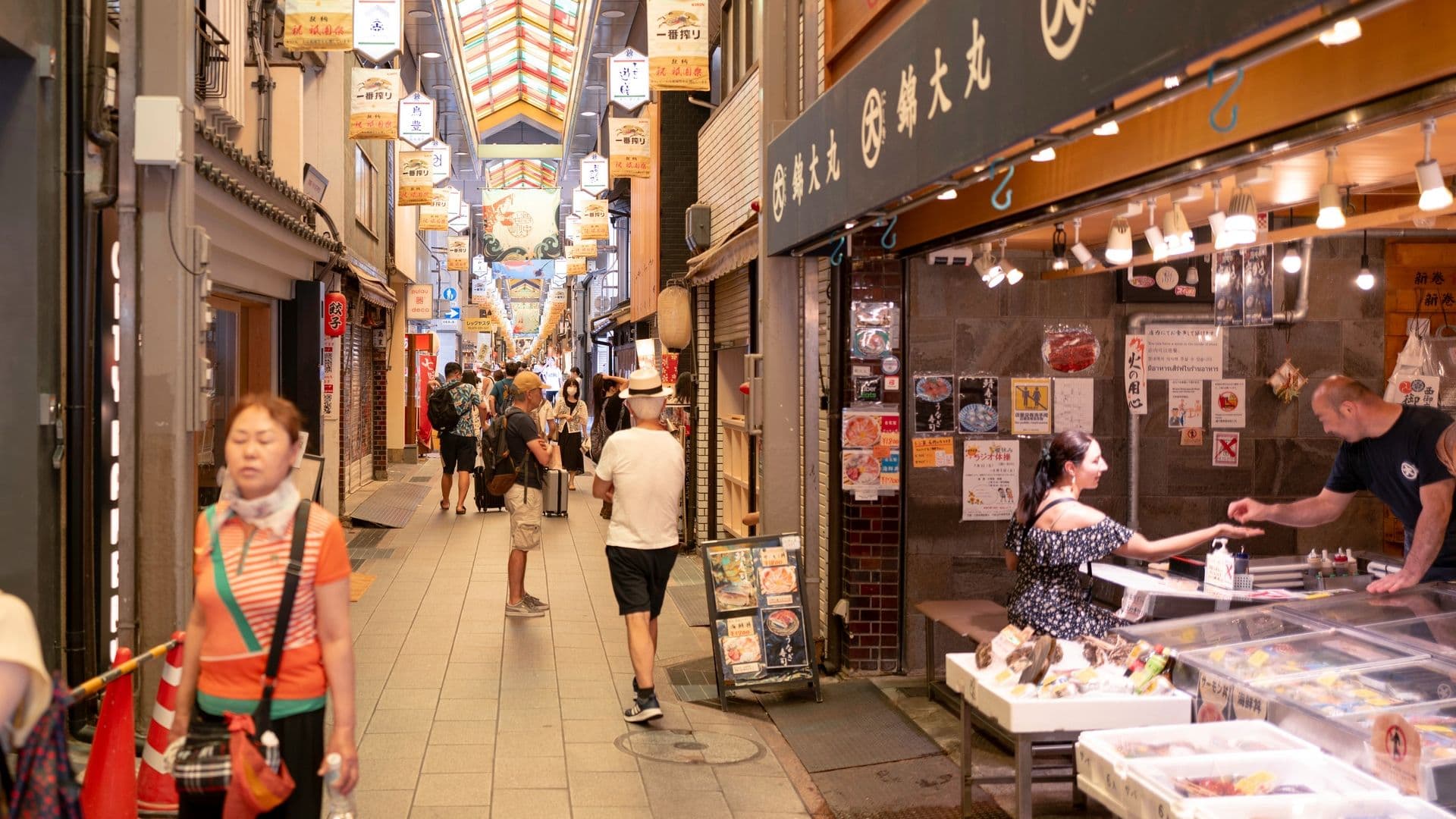 Indoor shopping alley with various shops, people walking, some wearing masks, and a seafood stall where a woman is purchasing from a man.