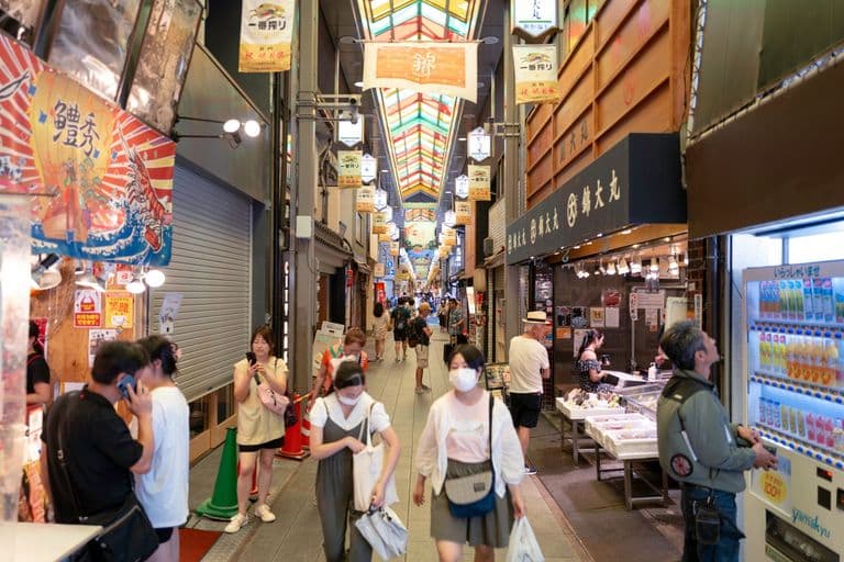 People walking and shopping in a covered Japanese shopping arcade with colorful banners and vending machines.