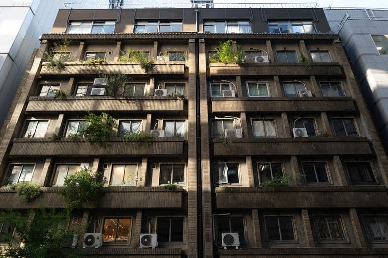 A multi-story apartment building with balconies and air conditioning units, some with plants and greenery, in an urban setting.