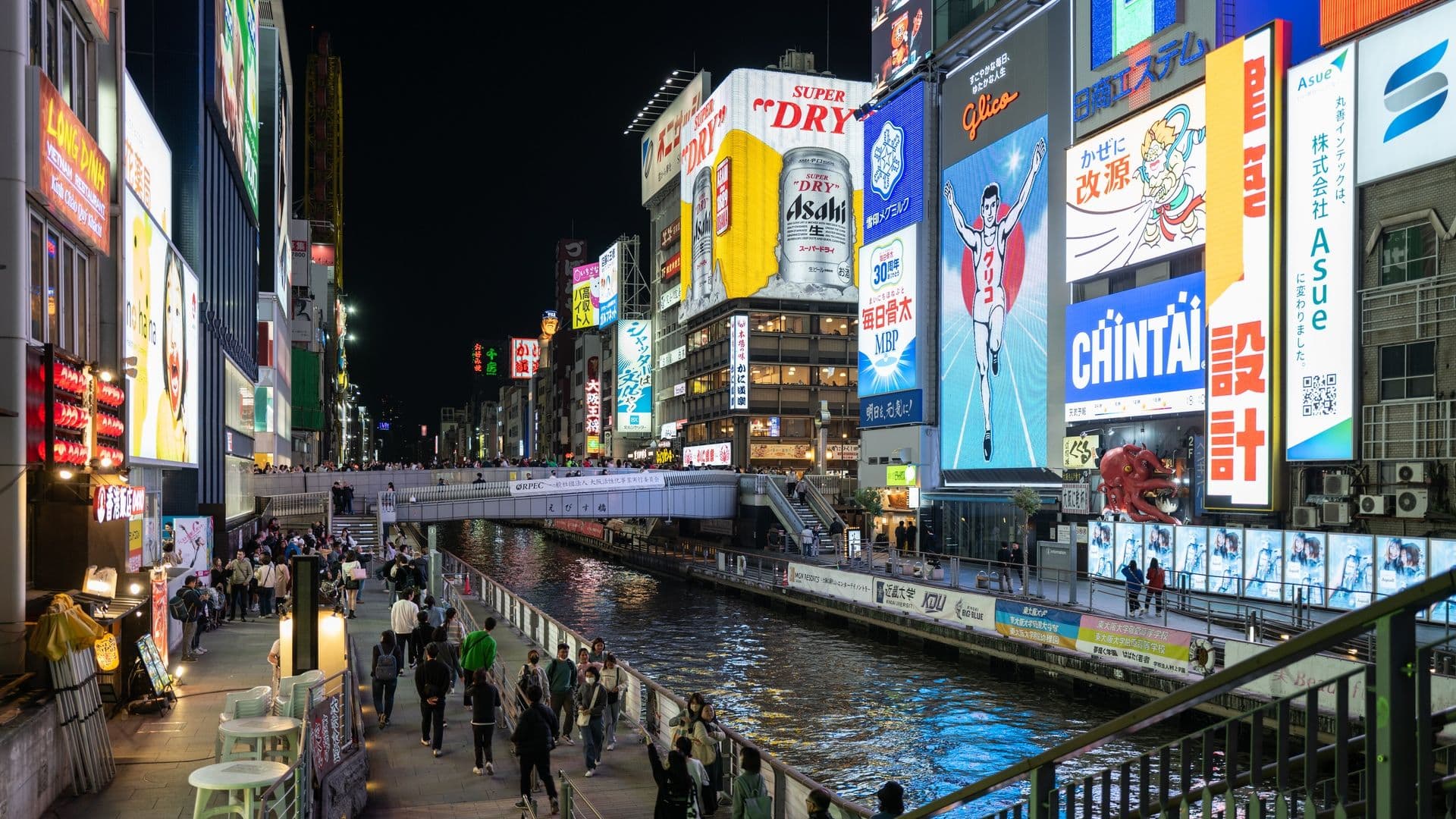 Night view of a busy commercial district with bright illuminated billboards, a canal, and people walking along the sidewalk.