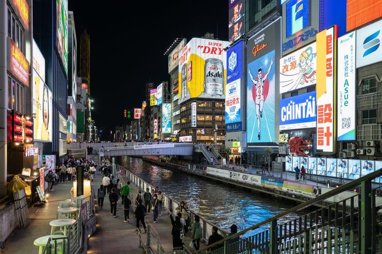 Nighttime scene of a brightly lit commercial district with skyscrapers, illuminated billboards, and advertisements along a canal. People are walking along the promenade beside the water.