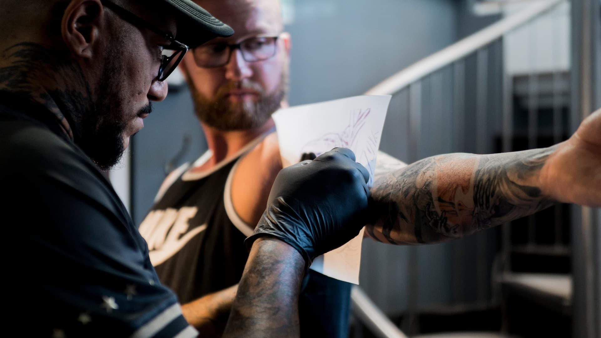 Tattoo artist working on a client's arm, with both focused on the tattoo, while the artist takes notes or makes sketches, in a tattoo studio setting.