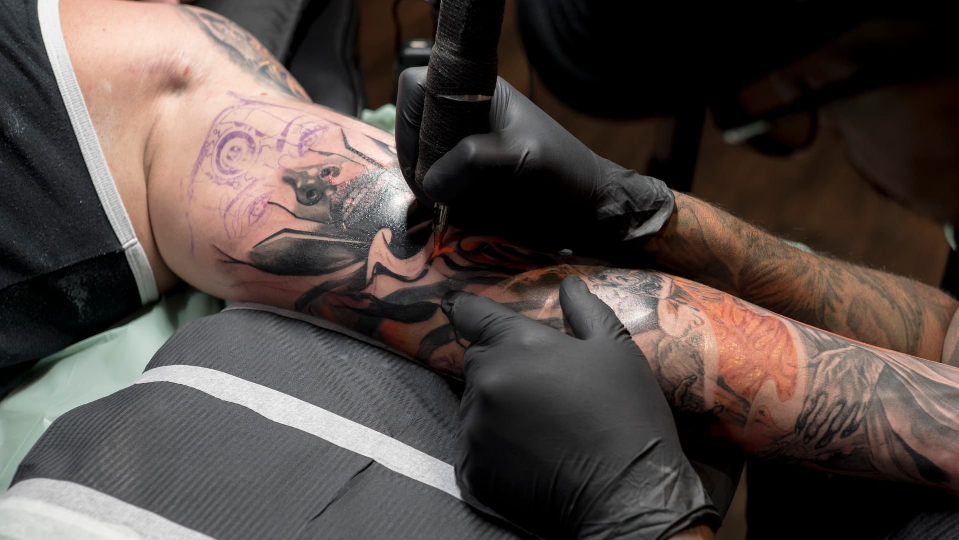 A person getting a tattoo on their upper arm from a tattoo artist wearing black gloves.