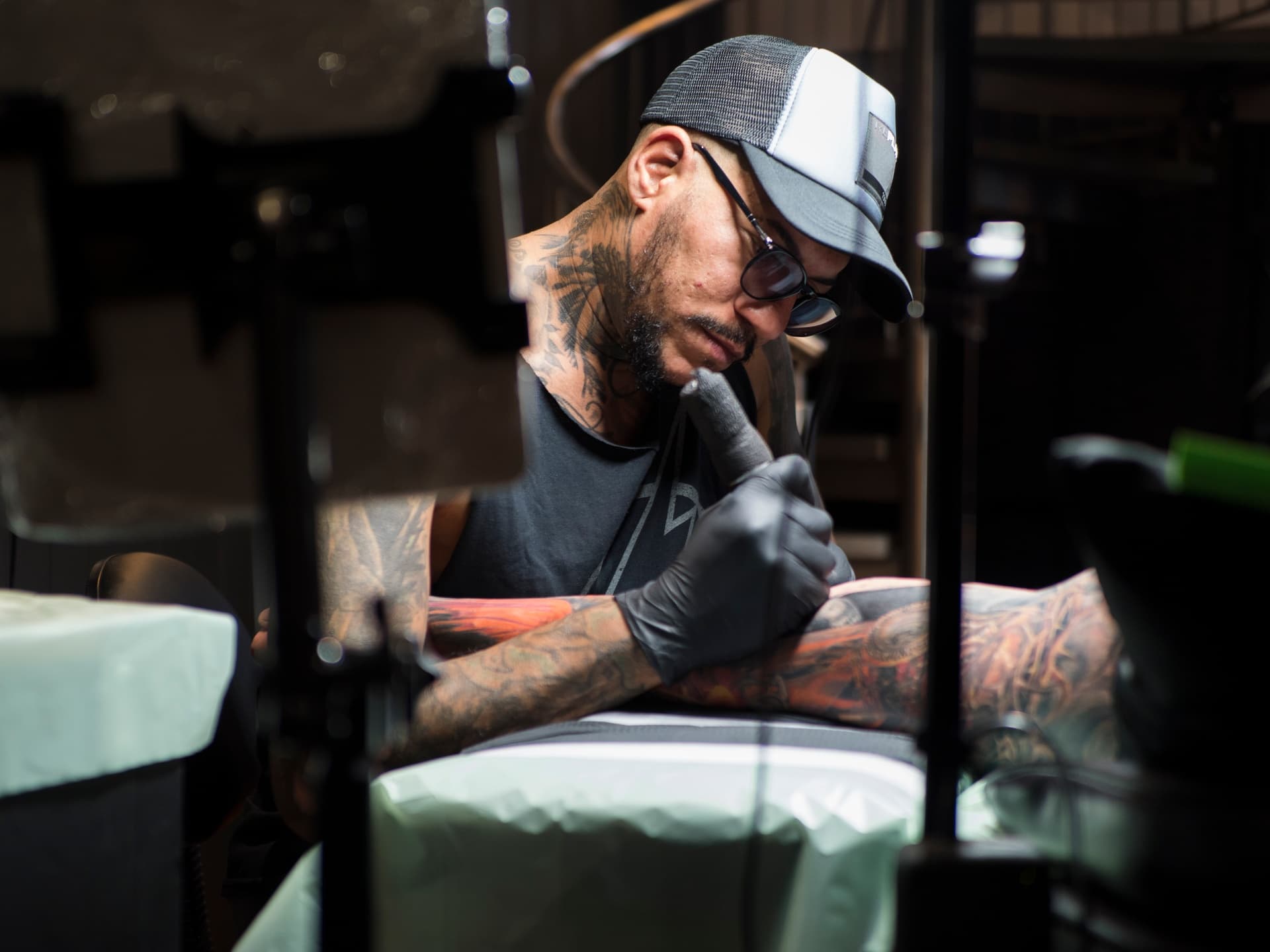 A tattooed man wearing glasses, a trucker hat, black gloves, and a sleeveless shirt, tattooing a person's arm in a tattoo studio.