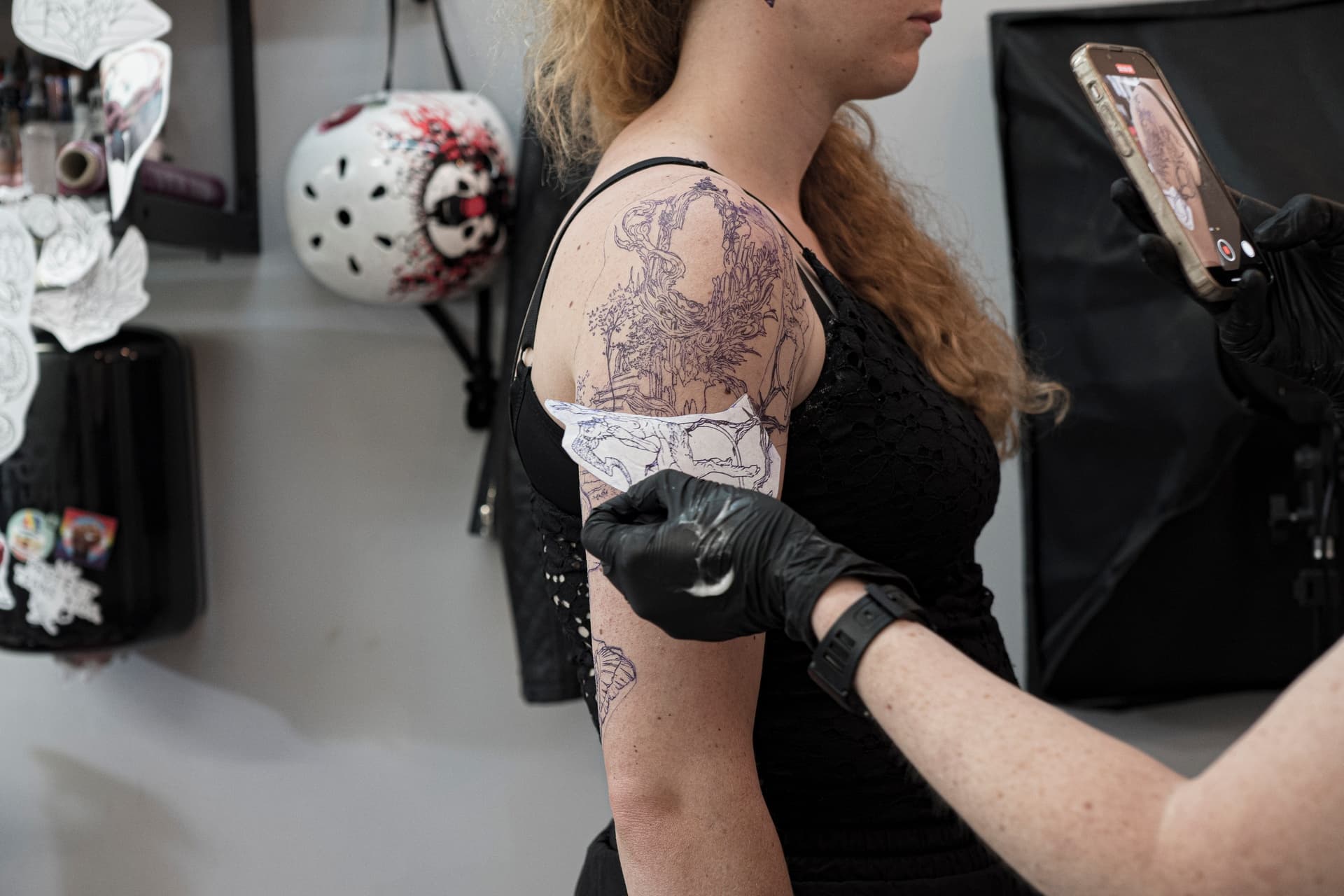 Woman getting tattooed on her upper arm by an artist wearing black gloves, with tattoo outlines visible on her shoulder and upper arm.