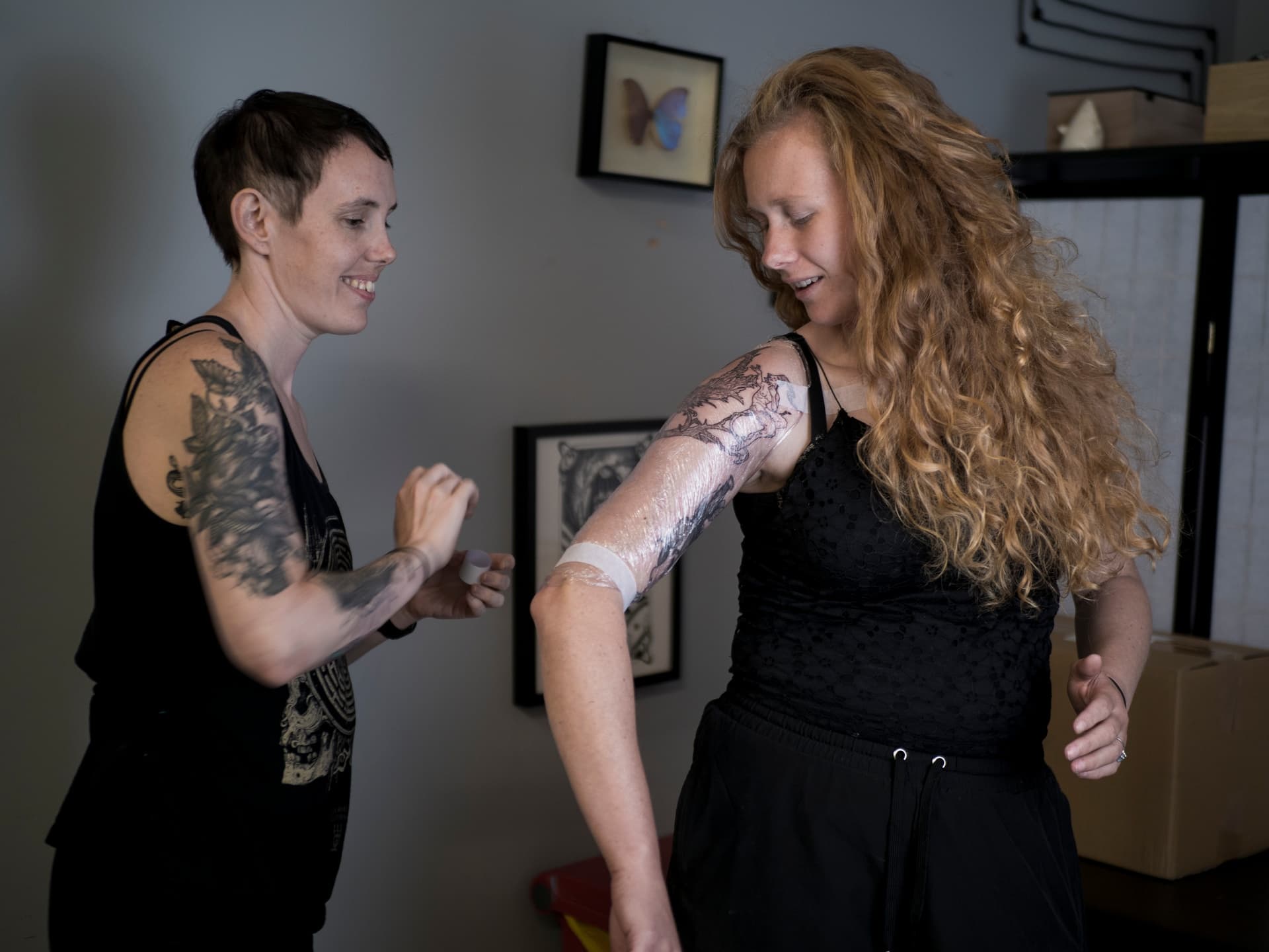 A woman with long red curly hair shows her tattooed arm to a smiling friend with short dark hair. The woman is in the process of getting a tattoo on her upper arm.