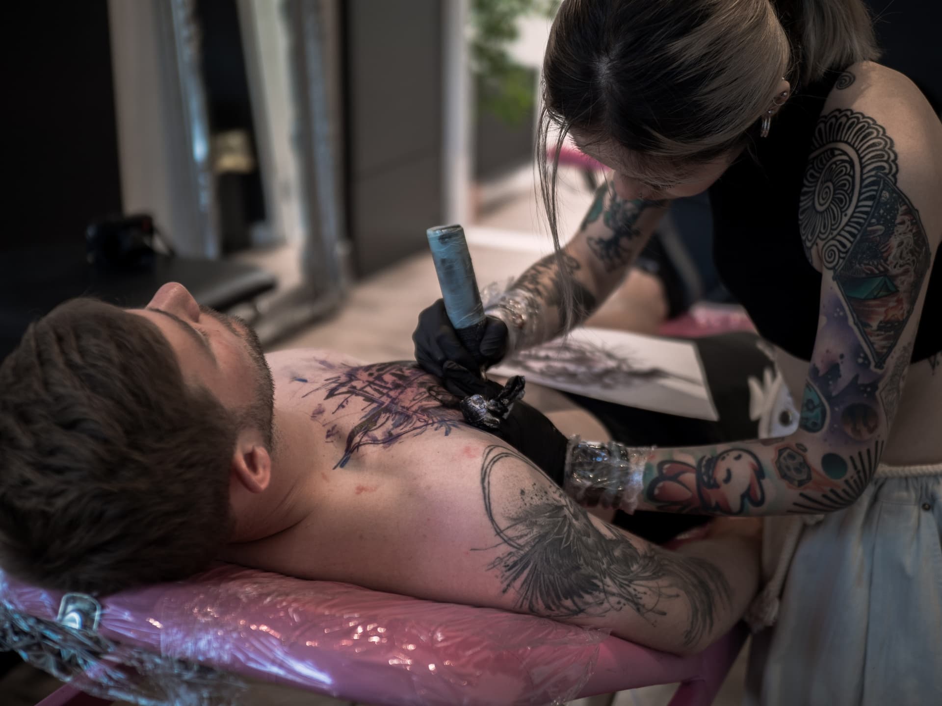 A woman with tattoos is tattooing a man with tattoos on his chest in a tattoo studio.