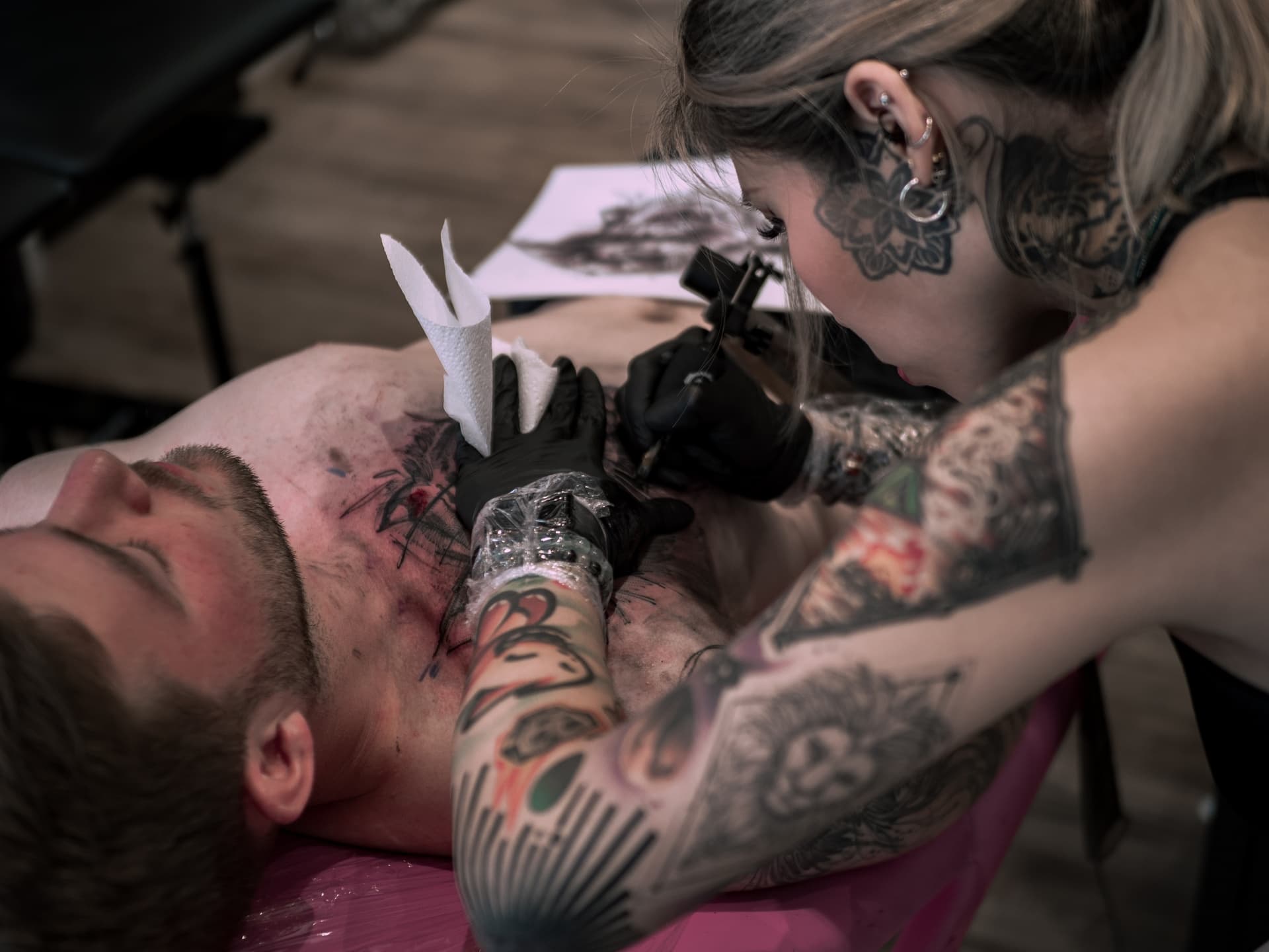 Tattoo artist with multiple tattoos ink tattoo on man's chest in tattoo studio.