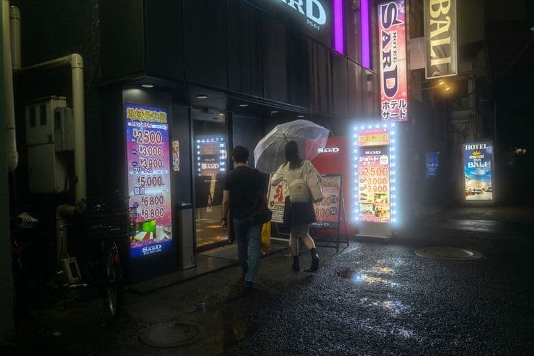 People walking into a nighttime nightclub or bar with bright, colorful signs in front and on the building, some rain puddles on the wet ground.