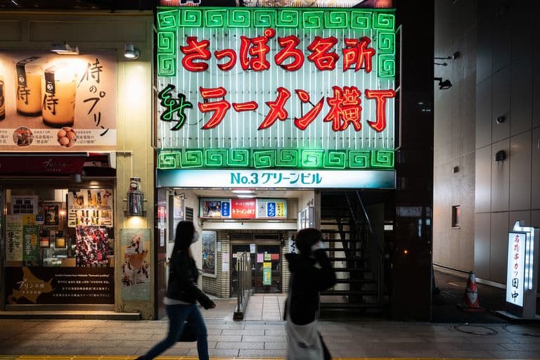 Neon sign in Japanese characters above an entrance to a building with stairs, with people walking past at night.