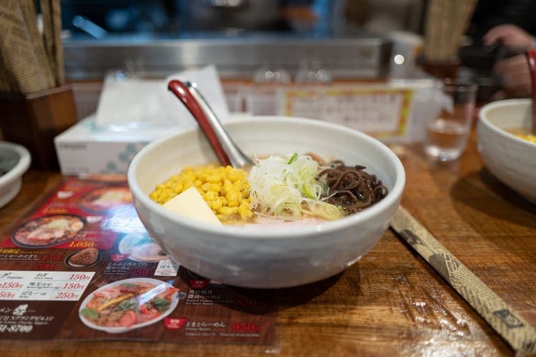 Bowl of ramen with corn, sliced green onions, brown mushrooms, a pat of butter, and a spoon, on a wooden table with a Japanese-style menu and chopsticks.