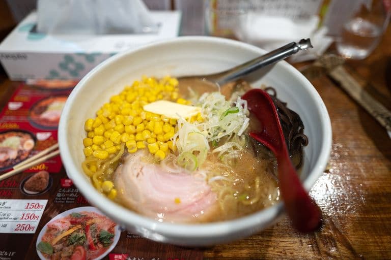 Bowl of ramen with corn, sliced green onions, pork, black noodles, and a pat of butter, on a wooden table with a red spoon and a metal spoon, and Japanese food menu in the background.