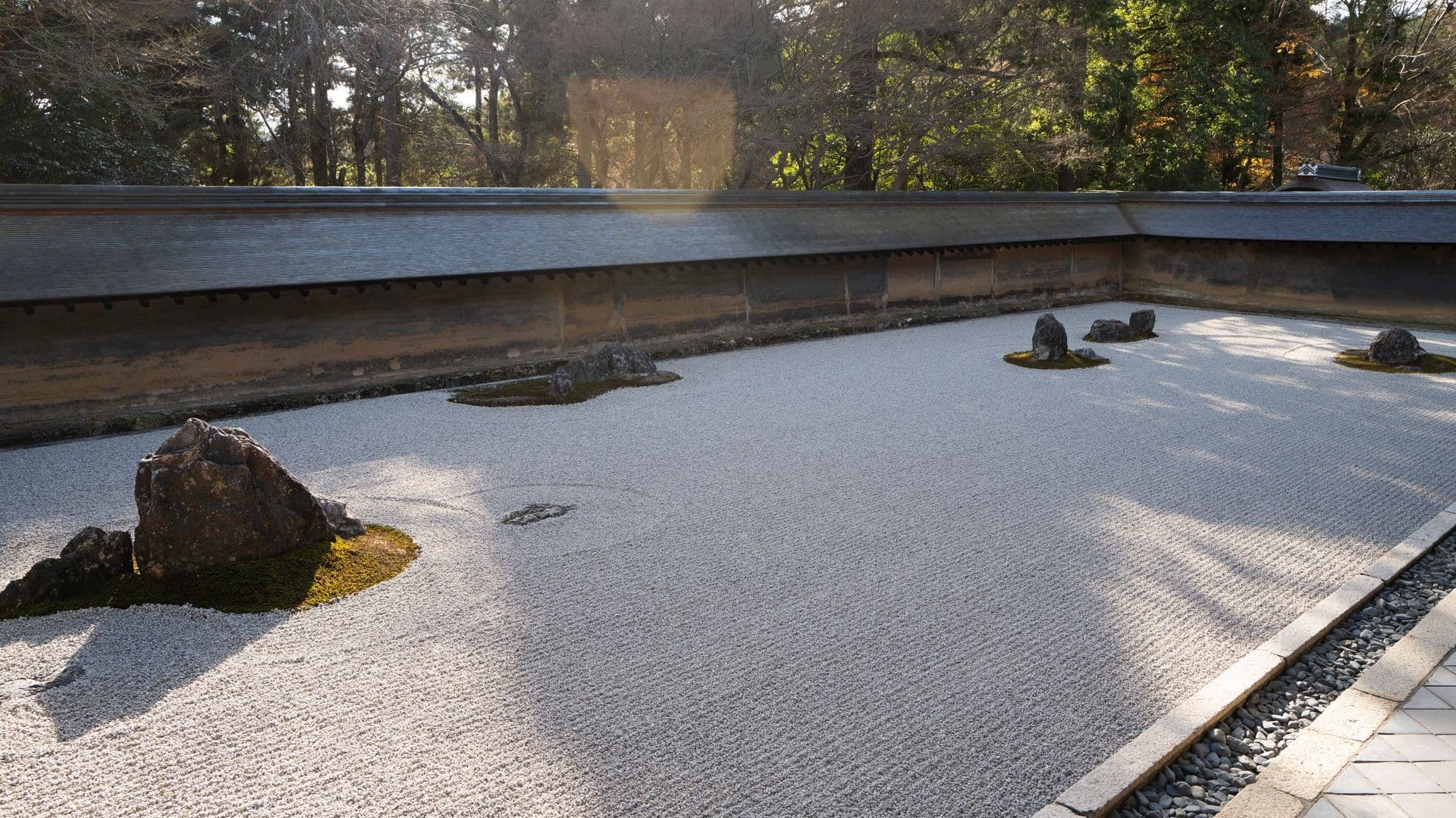 Traditional Japanese zen garden with white gravel, rocks, and moss patches, surrounded by trees.