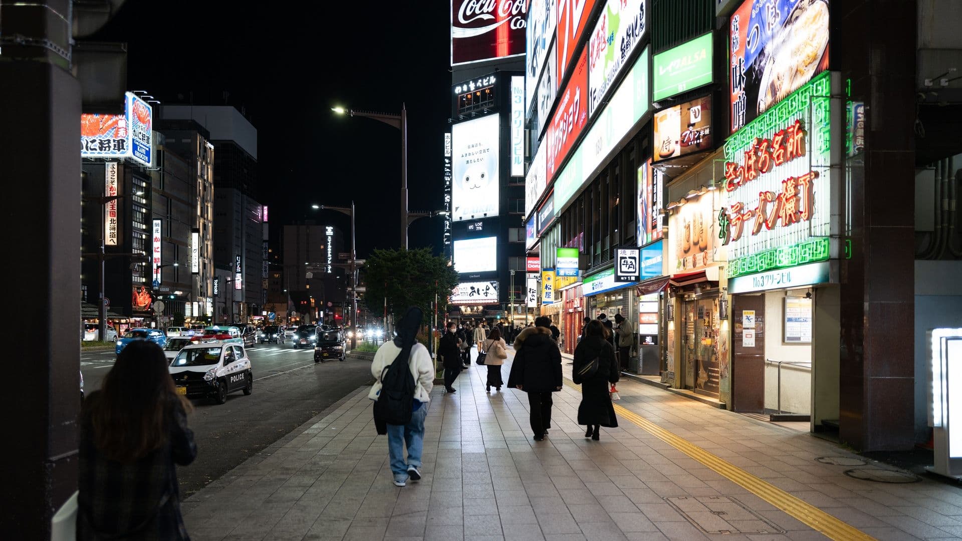 Nighttime city street scene with illuminated signs in Japanese, pedestrians walking on sidewalk, and cars on the road.